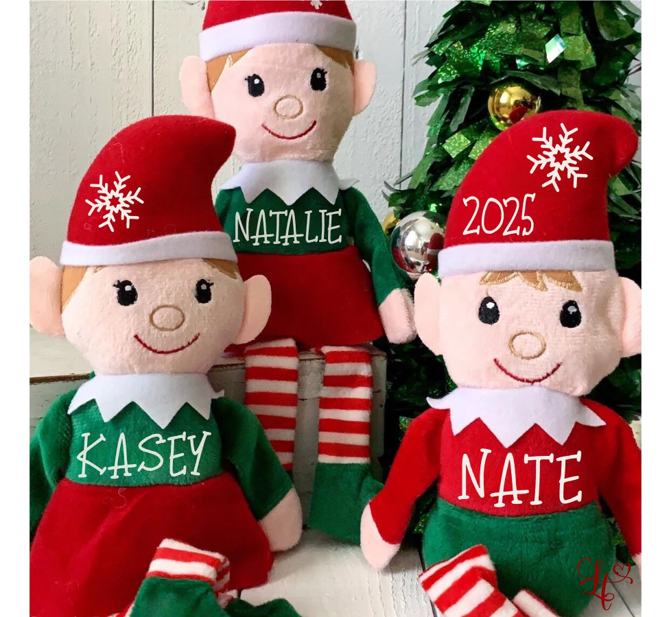 Three Christmas-themed elf dolls with names and the year on their clothing, standing in front of a decorated Christmas tree.