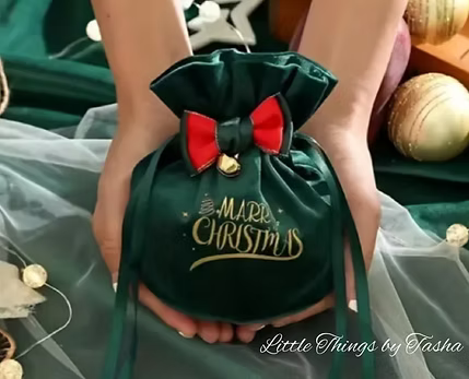 Green drawstring bag with a red bow held by hands, decorated with 'Merry Christmas' text.